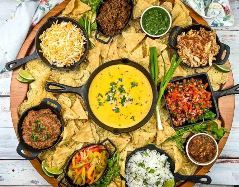 A platter with tortilla chips, queso dip, shredded cheese, cooked meats, salsa, pico de gallo, rice, vegetables, beans, and green sauce, arranged in small bowls.