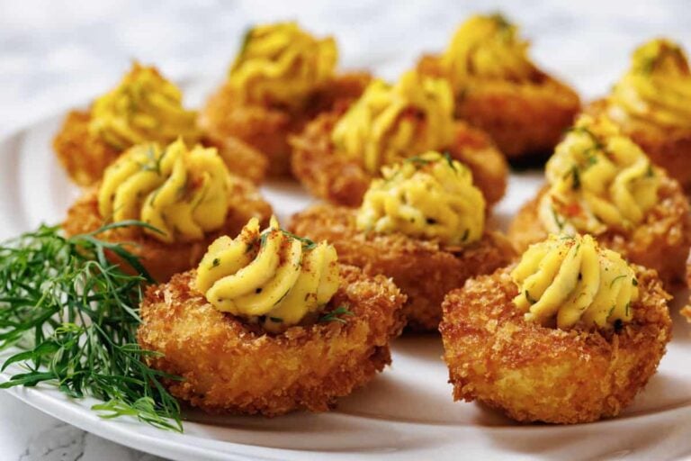 Close-up of breaded, bite-sized appetizers topped with piped yellow filling, arranged on a white plate with a garnish of fresh dill.