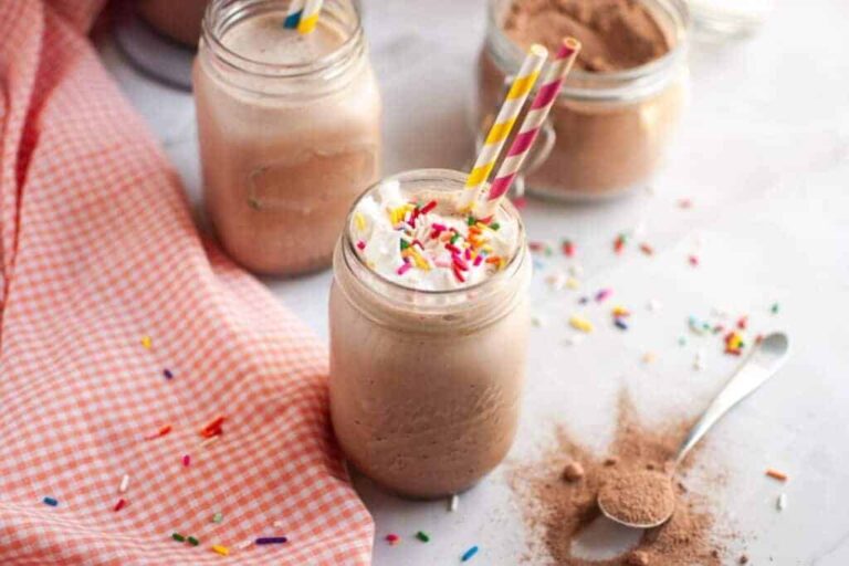 A mason jar filled with a chocolate milkshake topped with whipped cream and sprinkles, with two striped straws, sits on a table next to a spoonful of cocoa powder and a jar of mix.