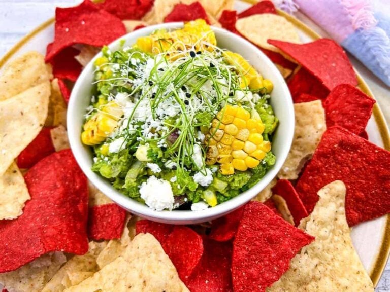 A bowl of guacamole topped with corn, cheese, and sprouts is surrounded by red and beige tortilla chips on a plate.