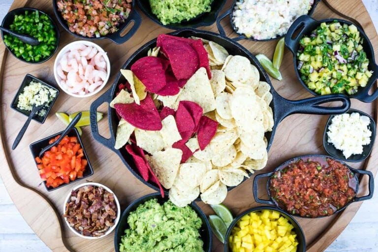 A wooden board with a skillet of tortilla chips, surrounded by small bowls of guacamole, salsa, pico de gallo, mango, shrimp, diced tomatoes, jalapeños, and other toppings.