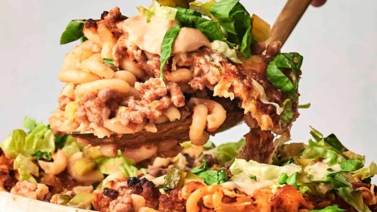 A wooden spoon lifts a portion of casserole with macaroni, ground meat, melted cheese, and fresh greens from a baking dish.