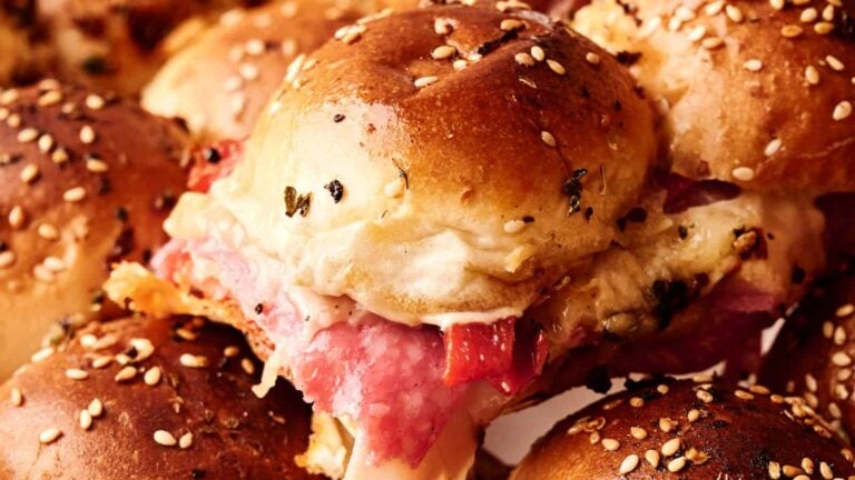 Close-up of a sandwich slider with sesame seed bun, layered with sliced meat and melted cheese, surrounded by similar sliders.