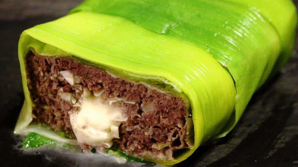 A slice of meatloaf wrapped in green leafy vegetable, filled with melted cheese in the center, sits on a dark surface.