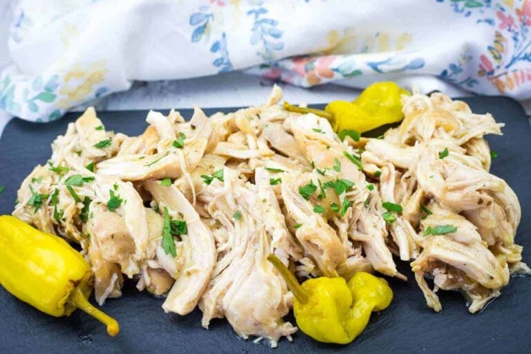 Shredded chicken garnished with chopped parsley, served on a black platter with whole pepperoncini peppers; a floral cloth is in the background.