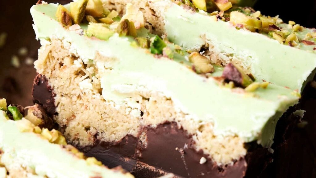 Close-up of sliced dessert bars with layers of chocolate, crumbly filling, and green topping, garnished with chopped pistachios.