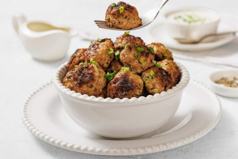 A white bowl filled with browned meatballs garnished with chopped herbs sits on a white plate; a fork holds one meatball above the bowl.