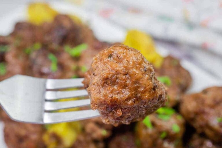 A close-up of a meatball held by a fork, with more meatballs and herbs visible in the blurred background.