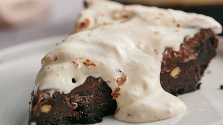A slice of chocolate brownie topped with a generous layer of vanilla cream sauce on a white plate.