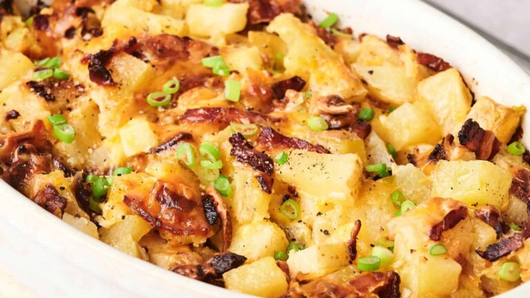Close-up of a baked casserole with diced potatoes, crispy bacon, melted cheese, and chopped green onions in a white dish.