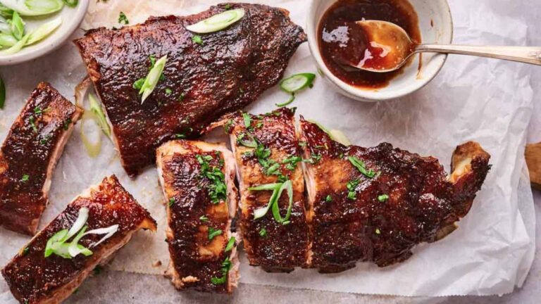 Sliced and whole barbecued ribs on parchment paper, garnished with green onions and herbs, with a bowl of barbecue sauce and a spoon beside them.