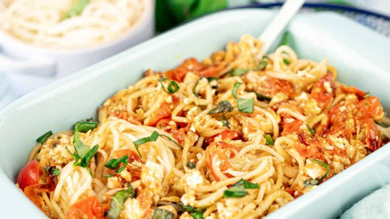A baked pasta dish with spaghetti, cherry tomatoes, crumbled cheese, and fresh basil, served in a white rectangular dish with a fork.