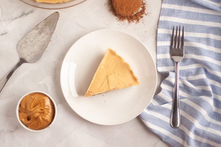 A slice of peanut butter pie on a white plate, with a fork, pie server, bowl of peanut butter, and blue-striped napkin on a marble surface.