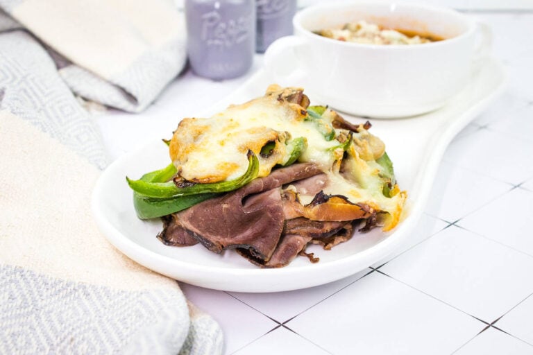 An open-faced sandwich with sliced roast beef, green bell peppers, mushrooms, and melted cheese on a white plate; a bowl of soup is beside it.