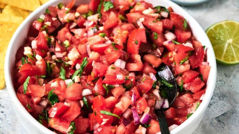 A bowl of fresh pico de gallo with chopped tomatoes, onions, cilantro, and jalapeños, with a spoon inside and tortilla chips and a lime nearby.