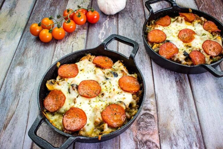 Two cast iron skillets with baked pepperoni and cheese dish on a wooden surface, with a cluster of tomatoes and a garlic bulb in the background.