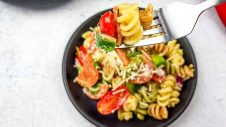 A fork holding rotini pasta with tomatoes, peppers, and shredded cheese above a plate of pasta salad.