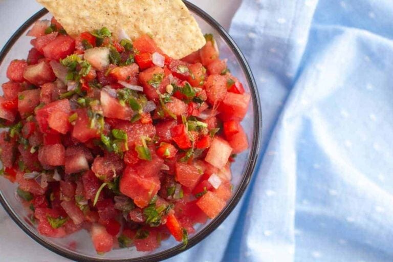 A bowl of watermelon salsa with diced vegetables and herbs, topped with a tortilla chip, next to a light blue cloth.