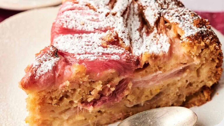 A close-up of a slice of rhubarb cake dusted with powdered sugar on a white plate with a spoon nearby.