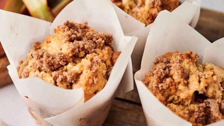 Three muffins with crumb topping are wrapped in white parchment paper and placed on a wooden surface.