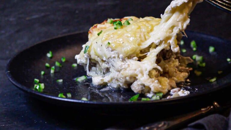 A serving of creamy, cheesy casserole is being lifted from a black plate, with melted cheese stretching and chopped green herbs scattered on the plate.