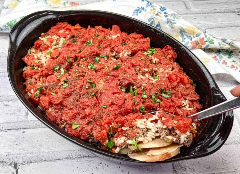A baked casserole dish filled with rice, ground meat, and tomato sauce, garnished with chopped herbs, with a serving spoon scooping out a portion.