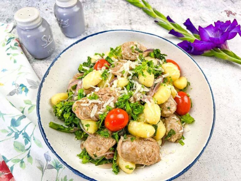 A bowl of gnocchi with sausage, cherry tomatoes, greens, and grated cheese, placed on a light surface with a floral napkin, salt and pepper shakers, and a purple flower nearby.