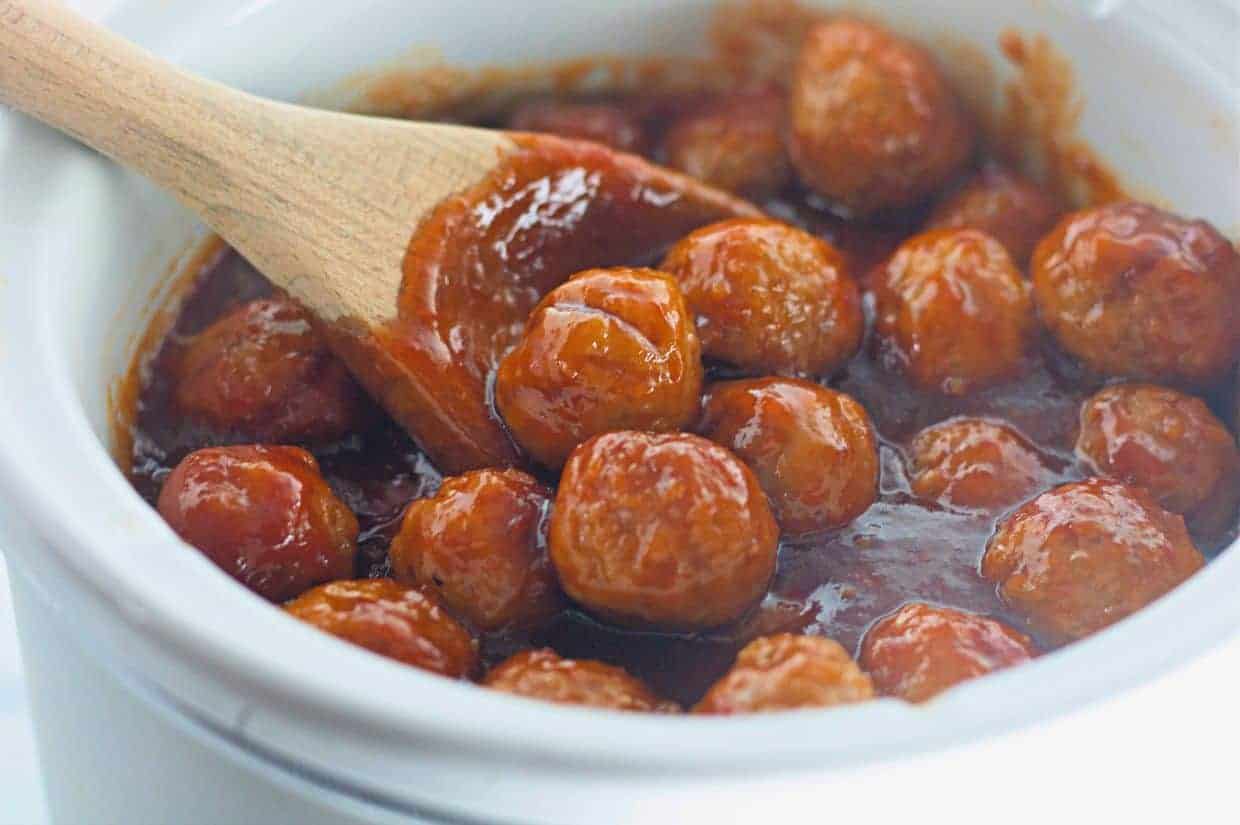 Honey-Garlic  Meatballs in a thick red sauce are being stirred with a wooden spoon in a white ceramic pot.