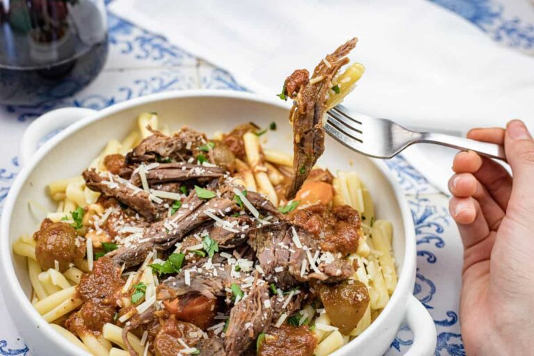 A hand holds a fork with shredded beef and pasta over a bowl of pasta topped with shredded beef, tomato sauce, and grated cheese.