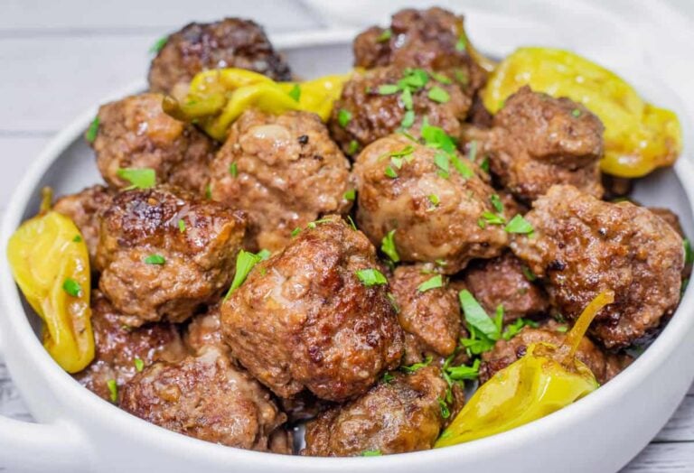 A white bowl filled with browned meatballs garnished with chopped herbs and yellow pepperoncini peppers.