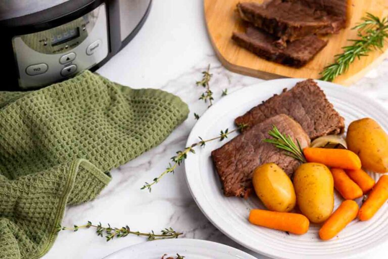 A plate with slices of beef, whole baby potatoes, and carrots, next to a slow cooker, a green kitchen towel, and herbs on a marble counter.