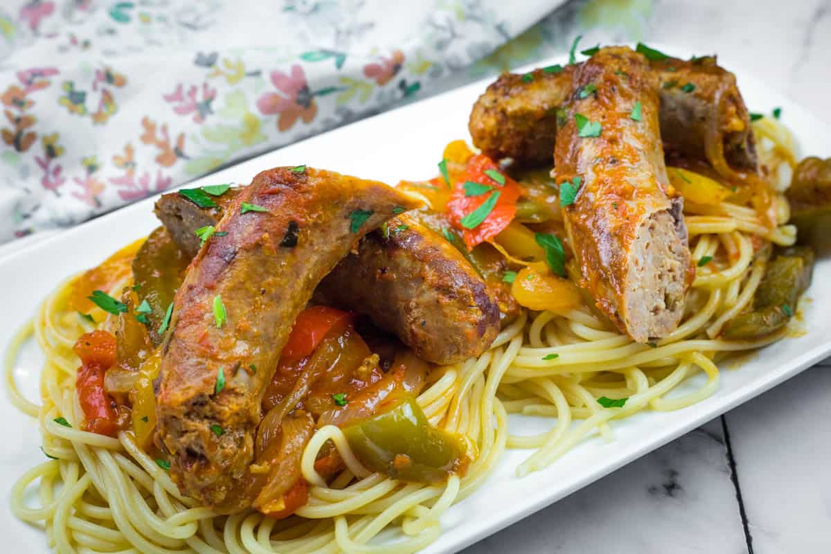 Slow Cooker Sausage & Peppers served over spaghetti garnished with herbs, on a white rectangular plate.