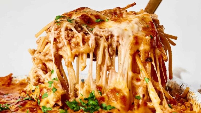 A close-up of a serving of baked spaghetti being lifted from a dish, with melted cheese stretching and garnished with chopped parsley.