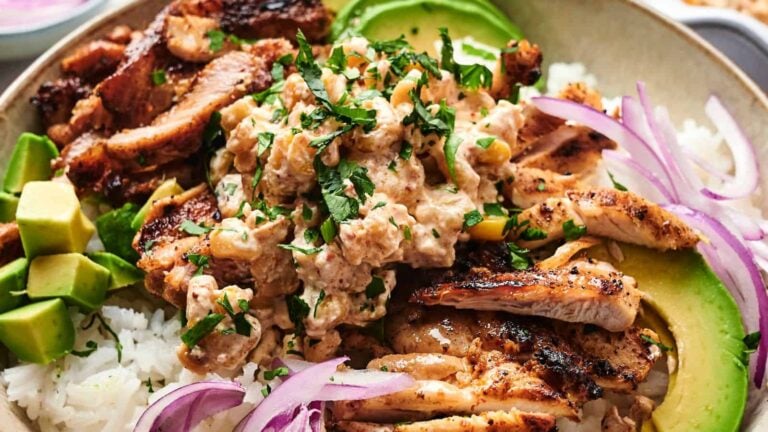 A bowl of white rice topped with grilled sliced chicken, creamy chicken salad, avocado chunks, sliced red onion, and chopped herbs.