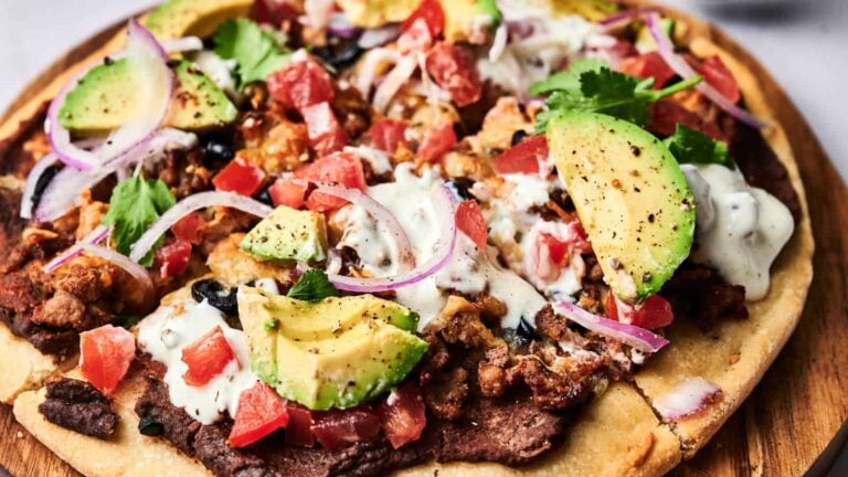 A pizza topped with black beans, ground meat, sliced avocado, diced tomatoes, red onion, cilantro, and a drizzle of white sauce on a wooden board.