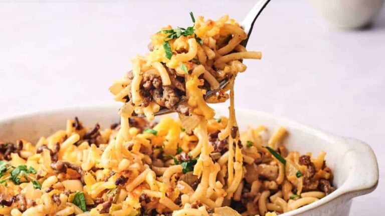 A fork lifts a cheesy ground beef and pasta casserole from a white baking dish, garnished with chopped parsley.