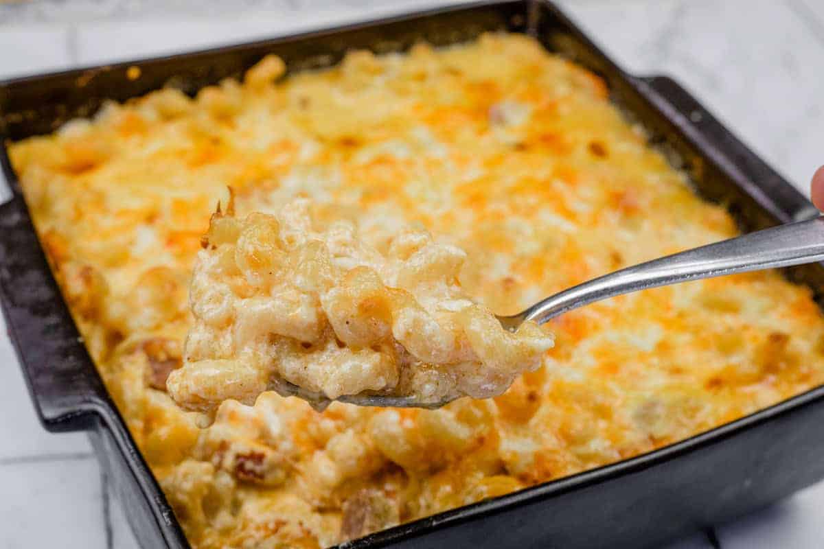 A spoonful of Tini’s Mac & Cheese with a Twist is held above a square baking dish filled with the same dish.