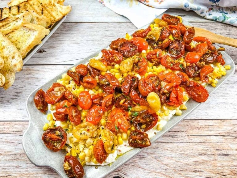 A platter with roasted cherry tomatoes, corn, and cheese, served on a rectangular tray with grilled bread on the side.