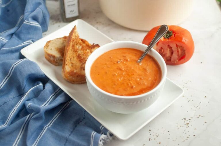 A white bowl of tomato soup with a spoon, served on a rectangular plate with two slices of toasted bread, a blue cloth, and a half-cut tomato beside it.