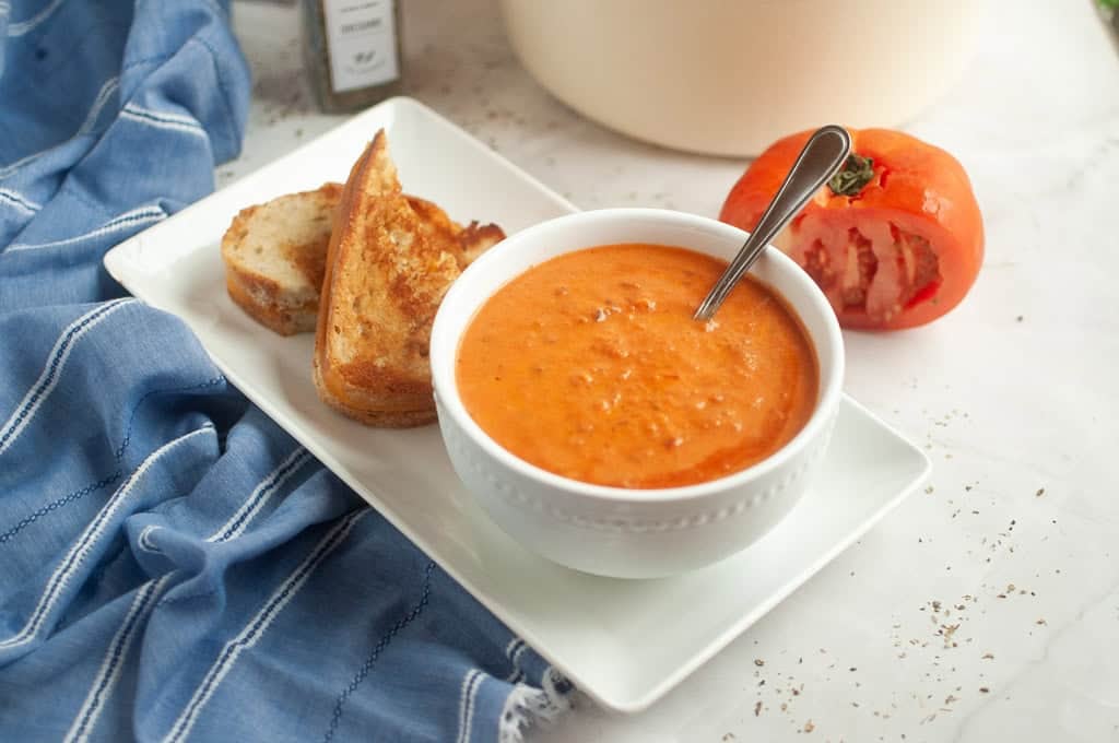 A bowl of tomato soup with a spoon, served with two slices of toasted bread on a white plate; a halved tomato and a blue cloth are nearby.
