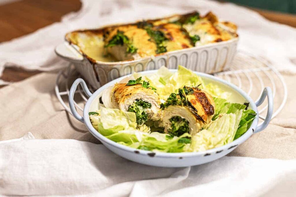 A serving dish with rolled chicken stuffed with green vegetables, placed on a bed of lettuce, with a baking dish of more stuffed chicken in the background.