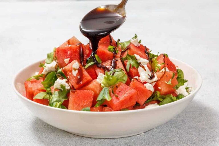 A bowl of watermelon salad with greens and feta cheese, drizzled with balsamic glaze from a spoon.