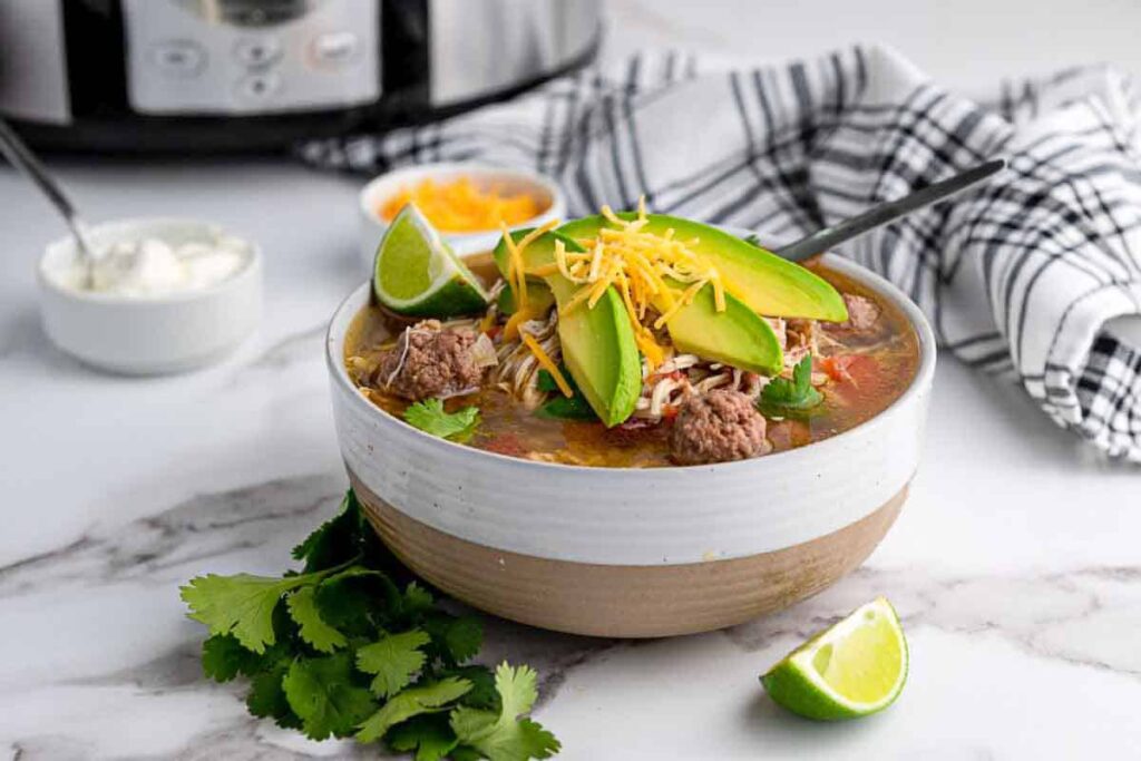 A bowl of beef soup topped with sliced avocado, shredded cheese, and cilantro, with a lime wedge, on a marble surface near a slow cooker, sour cream, and a checkered cloth.