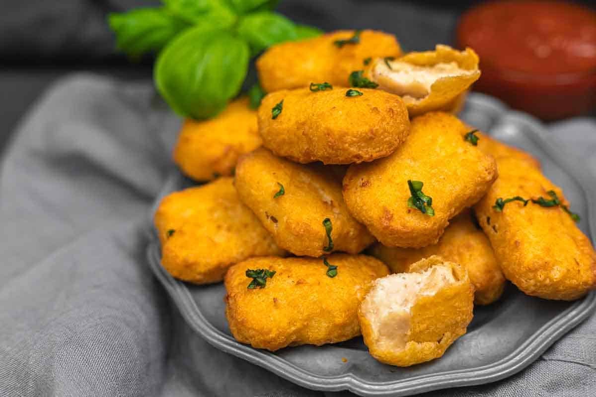 A plate with a pile of golden-brown, breaded chicken nuggets, garnished with small green herbs, on a gray cloth. Some nuggets are broken open, revealing the interior.
