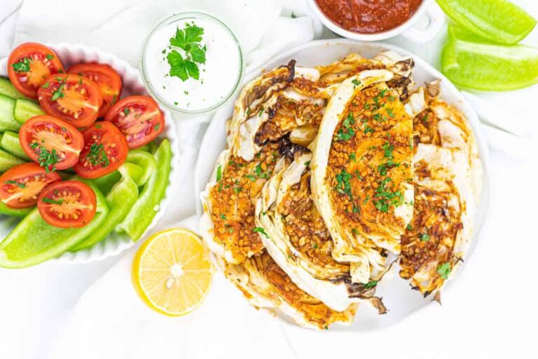 A plate of roasted cabbage wedges garnished with herbs, served with sliced green bell peppers, cherry tomatoes, lemon, and two dipping sauces.