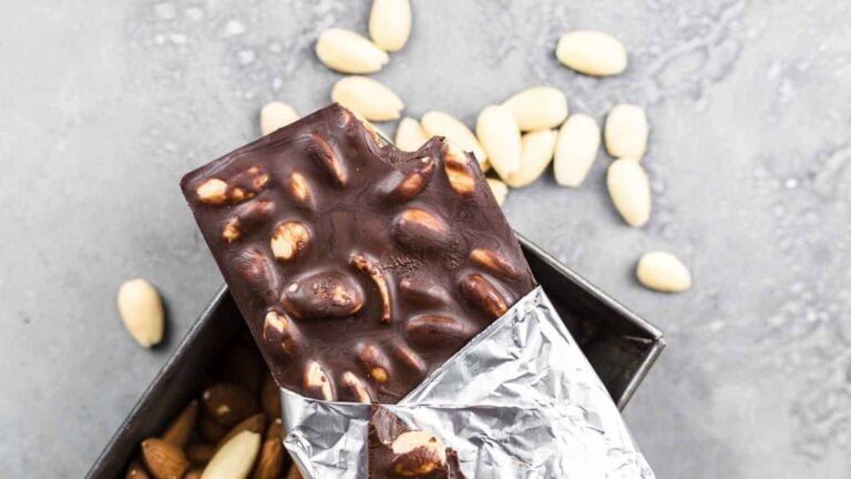 A partially unwrapped chocolate bar with whole almonds is shown on a gray surface, with scattered almonds in the background.