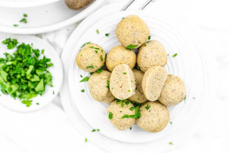A plate of round, beige matzo balls garnished with chopped parsley sits next to a small dish of additional parsley.