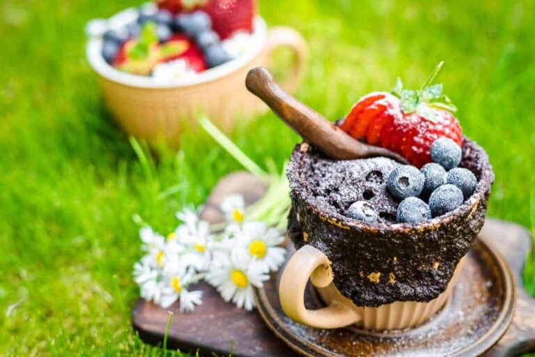 A chocolate mug cake topped with strawberries and blueberries sits on a saucer with daisies, on grass, with a second mug cake in the background.
