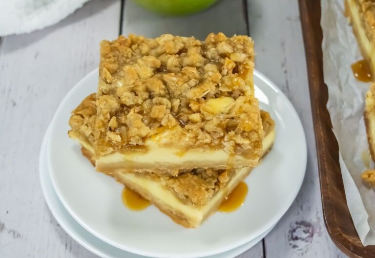 Two caramel-drizzled dessert bars with an oat crumb topping and creamy filling are stacked on a white plate, set on a light wooden surface.
