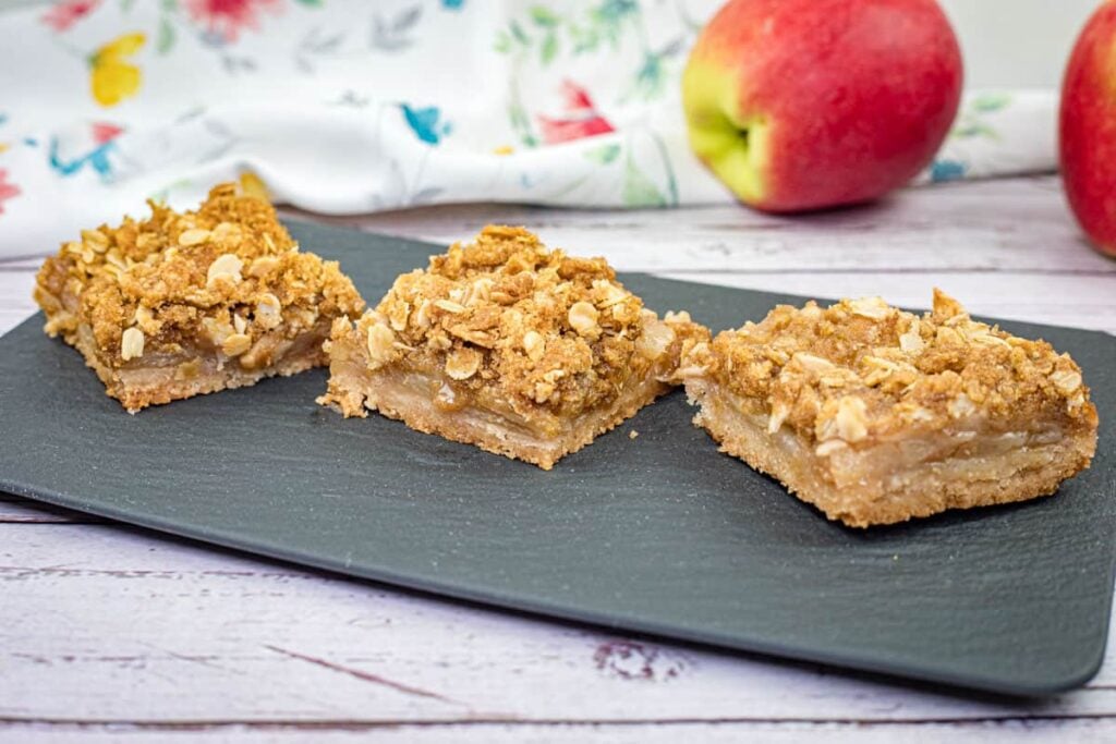 Three apple crumble bars with oat topping on a black serving tray, with apples and a floral cloth in the background.
